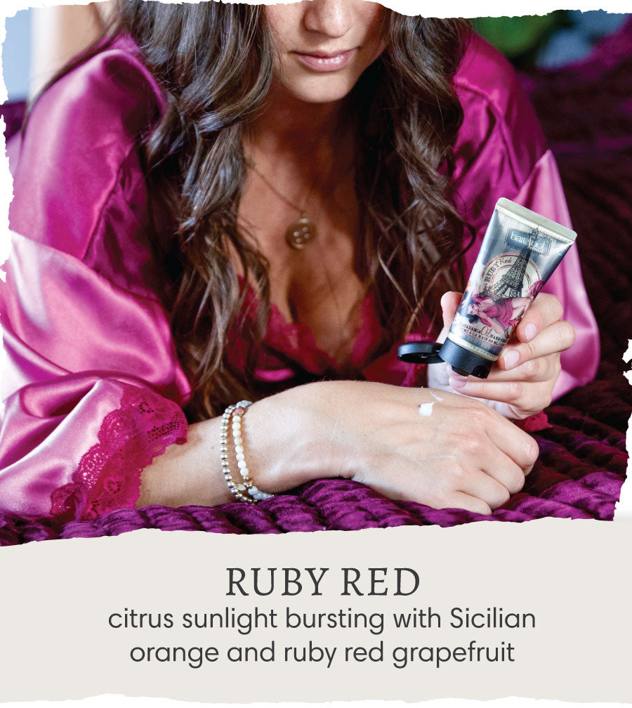 Woman in dark purple robe applying Barefoot Venus Ruby Red hand cream onto her hand with text overlay "Ruby Red - citrus sunlight bursting with Sicilian orange and ruby red grapefruit".