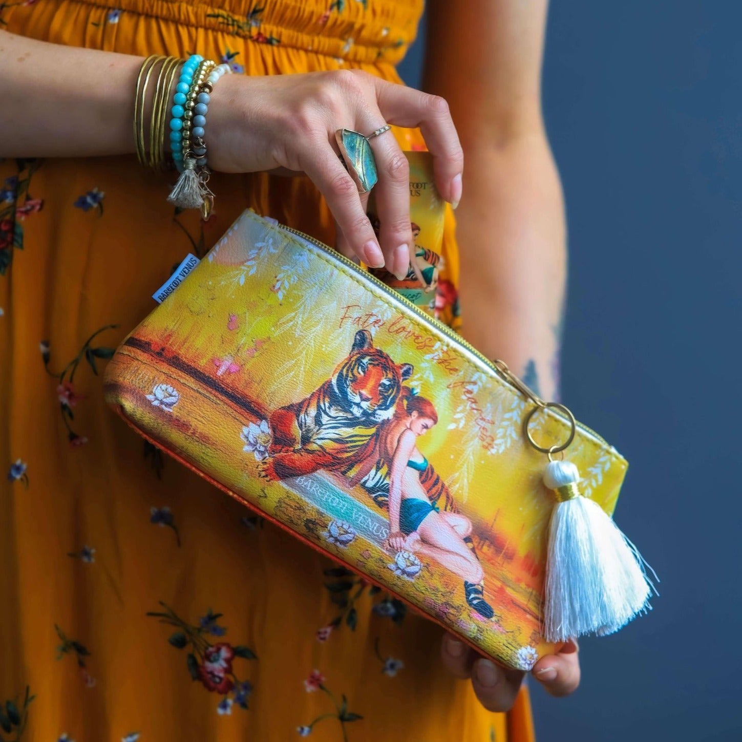 A yellow/orange pouch with a tiger and woman design featuring a white tassel, held by a person wearing a yellow dress.
