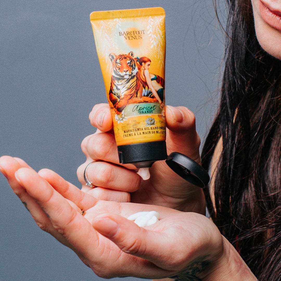 A person dispensing a tube of Apricot Brandy macadamia oil hand cream with a label featuring illustrations of a tiger and woman onto their hand.